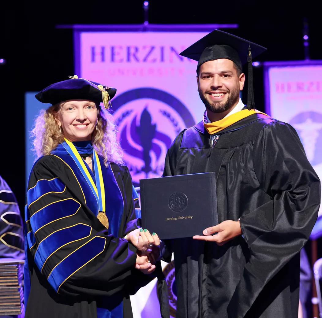 A proud graduate in a black cap and gown holding a Herzing University diploma, shaking hands with a smiling faculty member in academic regalia during a graduation ceremony, with university banners in the background