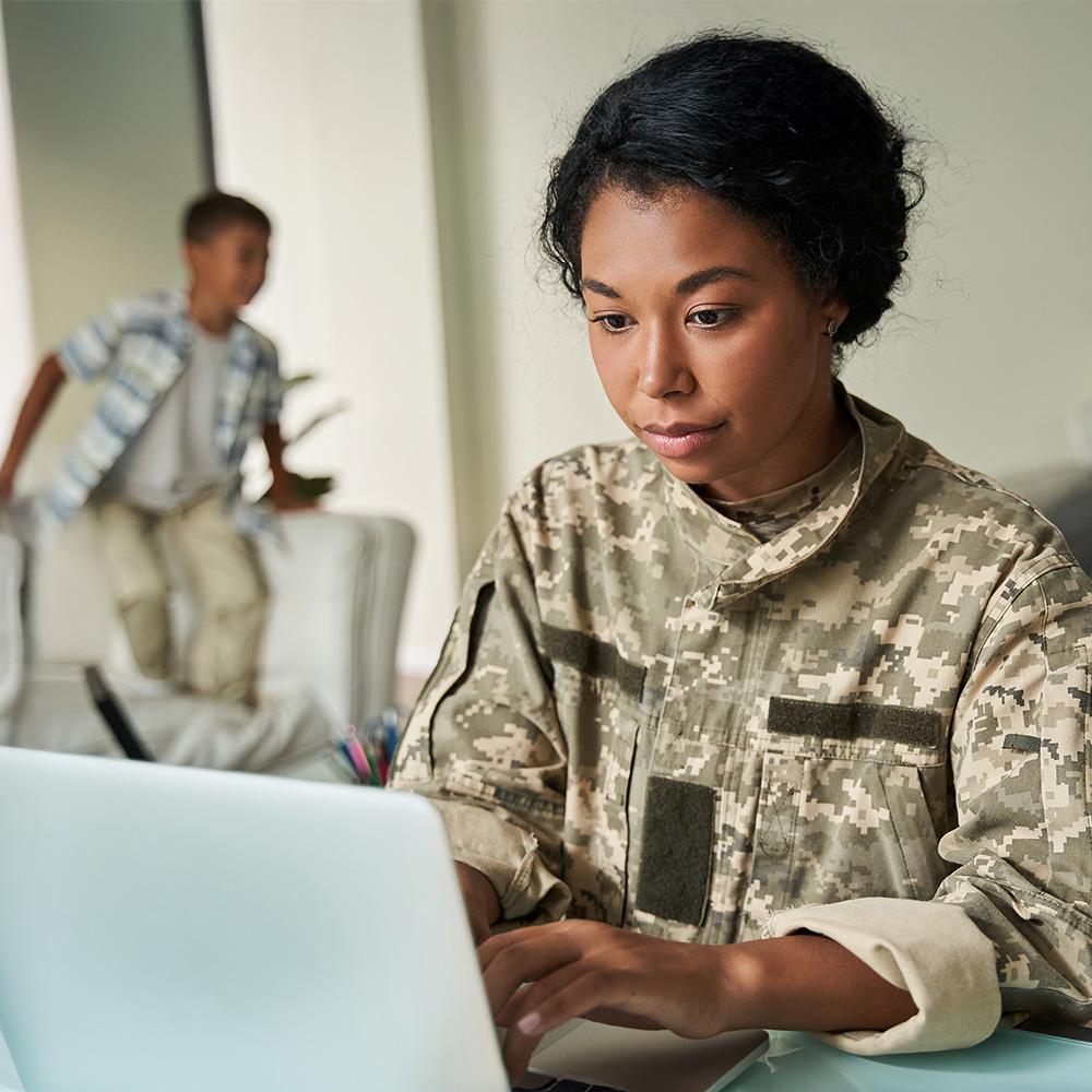 Military service member in camouflage uniform working on a laptop at home, balancing education and family responsibilities.